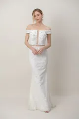 Vestido largo de novia de tul color blanco, con diseño de hombros descubiertos, mangas cortas caídas y corsé con apliques bordados a mano. Presenta una falda sirena con detalles florales bordados y cola.
