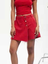 Short pollera de denim color rojo con diseño cruzado, cierre frontal de botones metálicos y acabado desmechado en el ruedo.