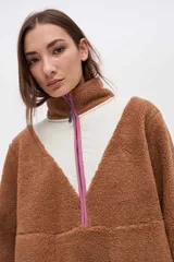 Buzo de sherpa color marrón con diseño de medio cierre frontal. Presenta un panel superior en forma de V en color beige y detalles de vivos en color fucsia en el cierre, cuello y bolsillos laterales. Cuenta con cuello alto y bolsillos frontales.