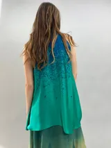 Blusa de satén con escote halter y cuello elevado anudado lateralmente. Presenta un diseño estampado degradado en tonos verde esmeralda y azul, con corte holgado y caída fluida.
