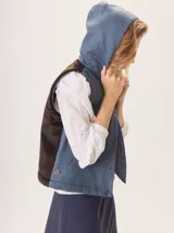 Chaleco con capucha, presenta una espalda de pelo sintético en color marrón oscuro con etiqueta de cuero negra y una capucha de tela tipo denim azul. Diseño original con mezcla de texturas.