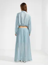 Blusa corta de tejido arrugado liviano, con cuello camisero, cierre frontal de botones y mangas largas abullonadas con puños. Presenta un ruedo elastizado que le otorga una silueta amplia y relajada.