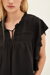 Camisa negra de viscosa con cuello redondo, cierre frontal con cordones y mangas cortas con volados. Presenta detalles de frunces en el pecho y hombros.