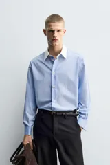 Camisa de corte regular fit confeccionada en algodón, con estampado de rayas verticales en tonos azul y blanco. Presenta cuello italiano en color blanco a contraste, cierre frontal de botones y mangas largas con puños dobles.