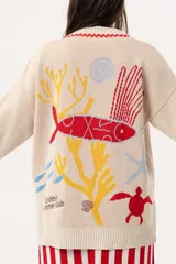 Saco tipo cárdigan oversize de tejido de punto color beige, con diseño inspirado en el universo marino que incluye ilustraciones de corales, peces y estrellas de mar. Presenta detalles de galón picot en color rojo en los bordes y puños, cierre frontal con botones rojos y un corte holgado.