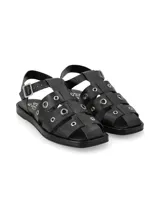 Sandalia tipo cangrejera de cuero graneado color negro, con diseño de tiras cruzadas decoradas con ojales metálicos, hebilla ajustable en el tobillo y suela plana.