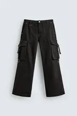 Pantalón cargo de corte relajado en color negro con efecto lavado. Presenta bolsillos laterales tipo cargo con solapas y cintas ajustables, bolsillos frontales y traseros, y detalles de argollas metálicas. Cierre frontal con cremallera y botón.