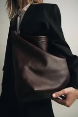 Bolso formato saca color marrón oscuro, confeccionado en piel. Tiene doble compartimento interior, asa de hombro y bolsillo interior con cierre mediante cremallera.