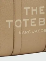 Cartera tipo tote de cuero granulado color tostado, con diseño estructurado y rectangular. Presenta doble asa superior fija y una correa larga ajustable y extraíble. En el frente, destaca el logo estampado con la inscripción 'THE TOTE BAG MARC JACOBS'.