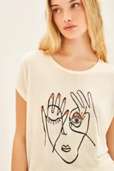 Remera de viscosa color crema, de calce suelto, escote redondo y manga corta. Presenta una estampa frontal centrada con el dibujo de un rostro y manos, y un bordado de un ojo con pedrería.