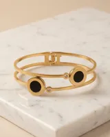 Esclava doble de acero con baño de oro, con dos círculos de esmalte negro y detalles de circonias. Cuenta con un sistema de apertura en la parte superior.