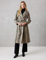 Trench de ecocuero con estampado animal print de serpiente en tonos beige, marrón y negro. Presenta cuello con solapa ancha, doble abotonadura frontal con botones negros, cinturón ajustable con hebilla metálica, mangas largas con presillas y botones en los puños, canesú suelto en la espalda, bolsillos laterales y forro interno.