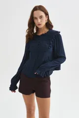 Blusa blanca de viscosa con cuello a la base, mangas largas abullonadas con puños ajustados, detalles de volados, encaje y botones frontales.