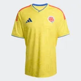 Camiseta de fútbol Adidas de la selección de Colombia, modelo Primera Equipación 26, color amarillo con un patrón sutil en relieve. Presenta cuello en V con ribete azul, logo de Adidas en azul y escudo de la federación colombiana. Las tres franjas de Adidas en los hombros son rojas y azules.