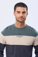 Remera de manga larga con cuello redondo y diseño de rayas horizontales en azul verdoso, beige y azul marino. Presenta una estampa frontal con texto y un parche decorativo en la manga.