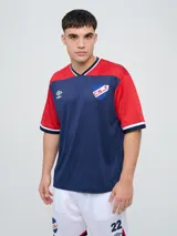 Remera de fútbol Umbro del Club Nacional de Football, de diseño tricolor con cuerpo azul marino, mangas y canesú rojos, y cuello en V con detalles en blanco y azul. Incluye el logo de Umbro en el pecho derecho y el escudo del club en el izquierdo.