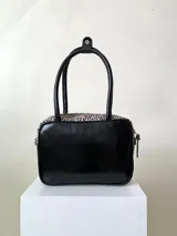 Cartera rígida negra con detalles en rojo y tachas plateadas. Tiene herrajes y cierre metálico plateado marca YKK, asa fina para llevar al hombro o en la mano y correa larga extra regulable. El interior es forrado, tiene un bolsillo con cierre enmarcado en cuero, y un segundo bolsillo plaqué.