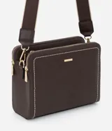 Cartera transversal de material sintético color marrón oscuro, de forma rectangular y estructura rígida. Presenta pespuntes decorativos en color blanco en los bordes frontales y cierres metálicos dorados. Incluye un asa ancha desmontable con diseño de rayas beige y marrón.