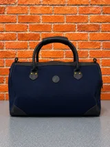 Bolso de viaje tipo duffle, confeccionado en lona azul marino con detalles en cuero negro en las asas, esquinas y bordes. Cuenta con un parche circular con logo en el centro y herrajes metálicos.