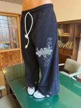 Pantalón de jogging negro de corte baggy, confeccionado en algodón. Presenta una estampa plastisol con bordado decorativo en la pierna izquierda, cintura elástica con cordón largo ajustable y botones en el ruedo para regular la botamanga.