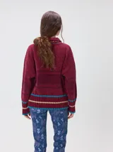 Pullover de punto con textura aterciopelada, diseño oversize, cuello polo con cierre frontal parcial y detalles de pespuntes en contraste. Presenta rayas horizontales en tonos tierra y un parche bordado con la letra M en el pecho.