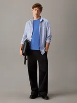 Camisa de manga larga Calvin Klein confeccionada en popelina de algodón elástico con diseño de rayas verticales. Presenta cuello en punta, cierre frontal de botones, bolsillo de parche en el pecho con logo bordado y bajo redondeado.