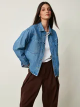 Camisa de denim celeste oversize con bolsillos frontales, cierre de botones metálicos y detalles de tachas en el cuello y la parte delantera.