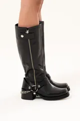 Bota estilo biker de cuero negro con caña alta y cierre metálico lateral. Presenta un diseño versátil con estribo y cadena desmontables, taco cuadrado y suela con plataforma.