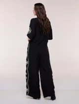Conjunto de dos piezas negro compuesto por camisa de manga corta con cuello camisero y pantalón palazzo de tiro alto. Presenta detalles de bordados florales en contraste a lo largo de los laterales del pantalón y en las mangas.