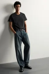 Jeans loose fit de hombre en denim de algodón color gris oscuro con efecto lavado. Presenta perneras con costuras giradas hacia los tobillos, tiro medio, cierre frontal con botones y bolsillos frontales con bolsillo cerillero.
