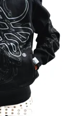 Hoodie con cierre frontal, color negro, con estampado gráfico de hojas de acanto en color blanco que recorre el frente y las mangas. Cuenta con bolsillos laterales y cierre metálico.