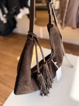 Cartera tipo hobo de textura gamuzada en color marrón oscuro, con diseño flexible y asa larga regulable. Presenta un detalle de flecos largos desmontables en uno de los laterales y cierre superior.