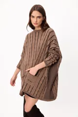 Sweater de punto con diseño de trenzas verticales, color crudo y camel, con bolsillos frontales y detalle de logo bordado. Presenta un corte holgado y mangas largas.