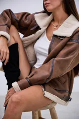 Campera tipo gamulán de corte oversize, confeccionada en cuero sintético marrón con efecto desgastado y forro interno de corderito sintético color crema. Presenta cuello solapa, cierre frontal con cremallera, detalles de tiras con hebillas en el cuello y en los laterales, y costuras reforzadas con bandas de cuero oscuro.