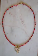 Collar con cadena de pequeños cristales cuadrados en tonos rojos y naranjas, y un dije en forma de hoja con baño de oro y circonitas transparentes.