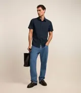 Camisa de manga corta con cuello inglés y cierre frontal abotonado, confeccionada en una mezcla de fibras de bambú, viscosa, poliéster y elastano. Presenta un diseño liso y entallado.