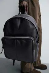 Mochila de cuero negro con compartimento principal y bolsillo frontal, ambos con cierre de cremallera. Incluye asa de mano superior, correas de hombro ajustables y parte posterior acolchada con banda de sujeción para equipaje.