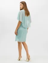 Vestido midi de tejido metalizado color turquesa, con sobrecapa de tul liviano sobre los hombros, detalle de pedrería en la cintura, volado lateral y tajo frontal.