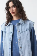 Chaleco de jean en tono celeste claro, con corte recto, cuello camisero, cierre frontal con botones metálicos y costuras visibles.