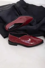 Mocasines de charol color bordó, de diseño vanguardista con corte asimétrico en el empeine, abertura en V, punta fina y suela plana con pequeño tacón.