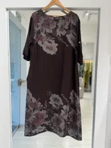 Vestido midi color marrón chocolate con estampa floral en tonos claros, escote en V y detalle de grandes botones circulares en el frente con tiras colgantes. Mangas cortas y corte holgado.