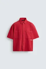 Camisa de algodón con estructura canvas, color rojo lavado, de corte relajado (relaxed fit) y manga corta. Posee cuello solapa, dos bolsillos de plastrón en el pecho y cierre frontal de botonadura. Presenta bordado y estampado de texto a contraste en el delantero.