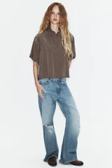 Camisa de corte fluido con cuello solapa y manga corta con dobladillo. Presenta un cierre frontal con botones ocultos bajo una tapeta y un diseño con bajo ligeramente más largo en la parte posterior.