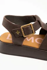 Sandalias estilo cangrejera de cuero marrón con plataforma, tiras cruzadas en el empeine, tira ajustable con hebilla metálica en el tobillo y detalle de estrella dorada grabada.