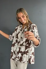 Blusa de corte holgado con estampado floral en tonos marrón y blanco. Presenta cuello camisero, cierre frontal con botones y un bolsillo en el pecho con detalle de lentejuelas. Las mangas son tres cuartos con trabas para ajustar.
