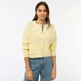 Cardigan de punto color amarillo suave, con cuello redondo, cierre frontal de botones y dos bolsillos de parche delanteros. Presenta mangas largas con puños acanalados y un corte recto.