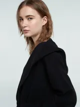 Abrigo largo de paño negro con capucha, cierre cruzado con lazo ajustable en la cintura y bolsillos delanteros con textura de pelo sintético. Presenta un diseño con espalda en nylon capitoneado.