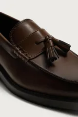 Mocasín de vestir color marrón oscuro, confeccionado en cuero, con detalle de antifaz y borlas decorativas en el empeine. Presenta una horma redonda y suela plana a tono.