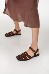 Sandalias planas de gamuza marrón, estilo pescador, con tiras entrelazadas, tachas doradas y tira ajustable con hebilla en el tobillo.