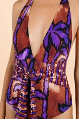 Traje de baño enterizo con diseño halter, escote profundo y estampado botánico en tonos púrpura y marrón cobrizo. Presenta una faja ancha fruncida en la cintura con un lazo decorativo central que cae hacia la parte inferior.